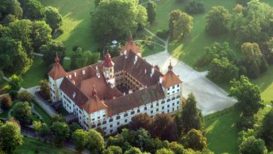 Das Bild zeigt das Schloss Eggenberg in Graz, Österreich, aus einer luftigen Perspektive. Das Schloss ist von großen, grünen Parkanlagen umgeben, die mit vielen Bäumen und Wiesen gestaltet sind. Die Architektur des Schlosses ist charakteristisch mit einem zentralen Gebäudeteil, der mehrere Giebel und Türme aufweist. Der Hauptbau hat eine helle Fassade mit dunklen Fensterrahmen sowie ein rotes Satteldach, das durch verschiedene Türmchen und Aufbauten ergänzt wird. Im Vordergrund erstreckt sich eine gepflasterte Fläche, die wahrscheinlich als Vorplatz dient. Die Umgebung scheint gepflegt und lädt zu Spaziergängen ein. Die gesamte Szenerie vermittelt ein Gefühl von historischer Bedeutung und natürlicher Schönheit.