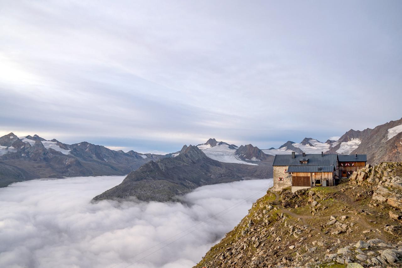 Eine Berghütte thront auf einem felsigen Grat über einem wolkenverhangenen Tal, umgeben von den schneebedeckten Gipfeln der Ötztaler Alpen.