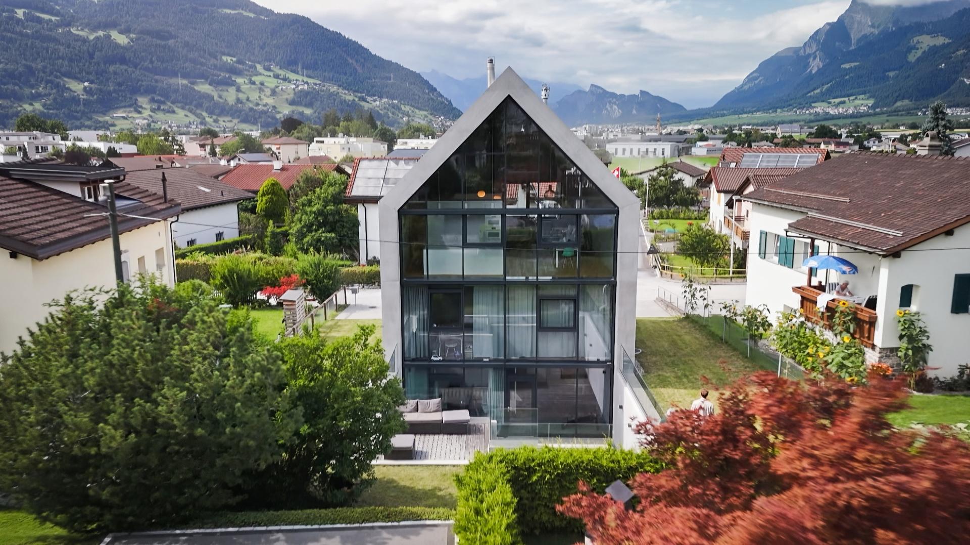 Das Bild zeigt ein modernes, mehrstöckiges Haus mit großen Glasfenstern. Es befindet sich in einer ländlichen Umgebung mit weiteren Wohngebäuden, Gärten und Bergen im Hintergrund.
