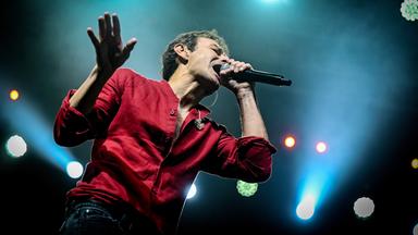 Untersicht eines Mannes im roten Hemd, der auf der Bühne steht und in ein Mikrofon singt, das er in einer Hand hält, während er die andere Hand anhebt.