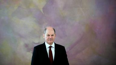 Ein Mann im Anzug steht mit dezentem Lächeln vor einer rosafarbenen Wand.