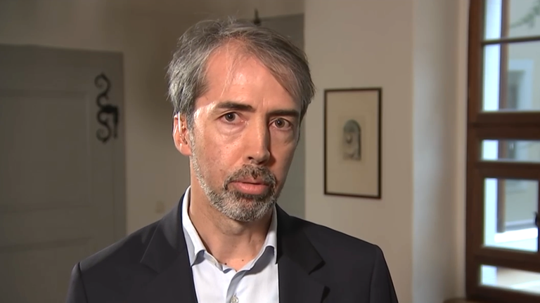 Ein Mann mit grauen Haaren und Anzug blickt in die Kamera. 