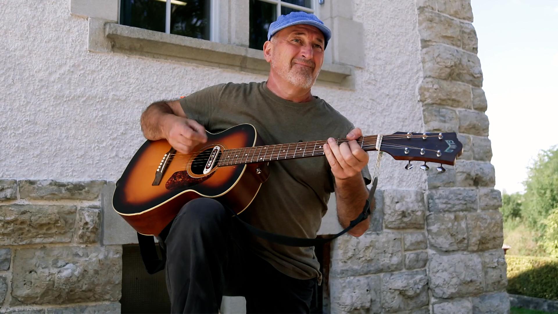 Ein Gitarre spielender Mann vor einem Haus sitzend.
