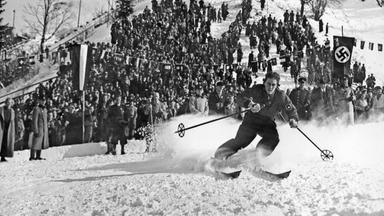 Historisches Schwarz-Weiß-Foto von einer Ski-Langläuferin, die vor einer Zuschauermenge den Berg hinunter saust. Am rechten Rand des Bildes ist eine Hakenkreuz-Flagge zu sehen.