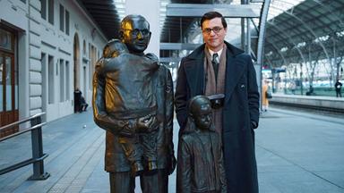 Johnny Flynn in seiner Rolle als junger Nicholas Winton steht mit einem Fotoapparat um den Hals auf einem Bahngleis neben einer Statue von Nicholas Winton. Die Skulptur besteht aus drei Personen: Nicholas Winton trägt einen Jungen auf dem Arm, von dem nur der Rücken zu sehen ist, neben ihm steht ein Mädchen.