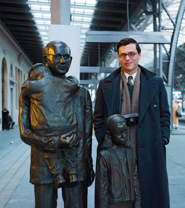 Johnny Flynn in seiner Rolle als junger Nicholas Winton steht mit einem Fotoapparat um den Hals auf einem Bahngleis neben einer Statue von Nicholas Winton. Die Skulptur besteht aus drei Personen: Nicholas Winton trägt einen Jungen auf dem Arm, von dem nur der Rücken zu sehen ist, neben ihm steht ein Mädchen.
