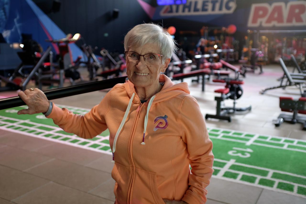 Eine ältere Dame mit kurzen, grauen Haaren und mit einer orangenen Kapuzenjacke bekleidet steht in Raum mit vielen Sportgeräten.