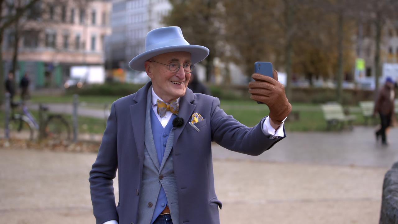 Ein älterer Herr mit Hut, Weste und Fliege steht auf einer Straße und hält ein Handy in der Hand, um ein Selfie zu machen.