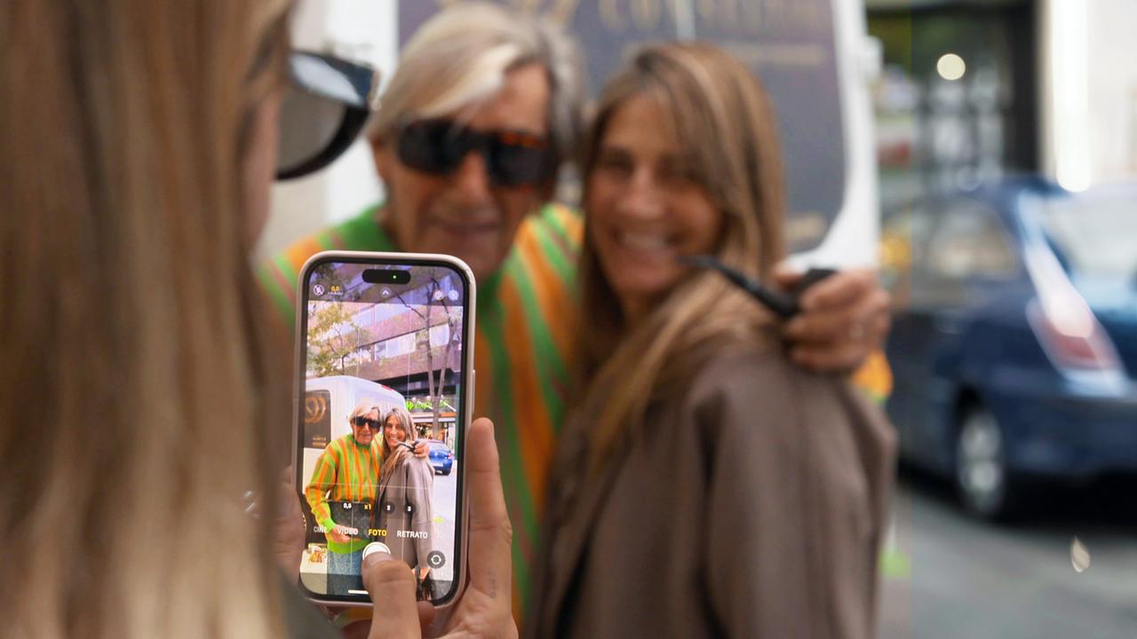 Fotosession mit einem Handy. Der leuchtende Handy-Bildschirm zeigt einen, älteren Mann mit Sonnenbrille, neben ihm eine jüngere Frau mit langen Haaren. Im Hintergrund die beiden, die gerade fotografiert werden, noch einmal unscharf zu sehen.