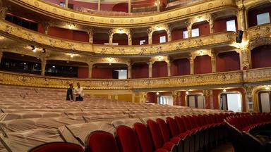Das Bild zeigt den Innenraum eines Theaters. Im Vordergrund sind Sitzreihen aus gepolstertem Stoff zu sehen, die teilweise mit transparentem Plastik abgedeckt sind. Zwei Männer stehen im Bild; einer von ihnen hat einen kurzen Bart und trägt ein dunkelgrünes Hemd, der andere trägt ein weißes Hemd und hat kurz geschnittene Haare. Der Mann im weißen Hemd zeigt mit der Hand in eine Richtung, während der andere ihn aufmerksam anschaut.   Die Wände des Theaters sind reich verziert mit goldenen Ornamenten und Skulpturen. Die Balustrade, die die Sitzreihen umfasst, hebt sich durch ihren filigranen Kunststil hervor. Deckenlichter sind sichtbar, und der Raum wirkt gut beleuchtet, während im Hintergrund große Fenster zu erkennen sind. Es handelt sich um einen eleganten und historischen Raum, der für Aufführungen konzipiert ist.