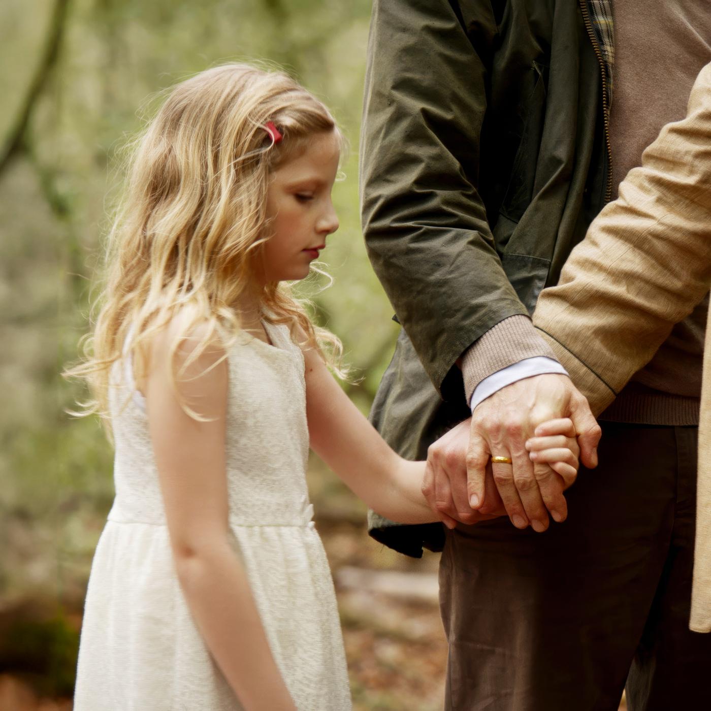 Die kleine Lucy (Carlotta von Falkenhayn) steht mit abgesenktem Blick in einem weißen Kleid im Wald und hält die Hand eines korpulenten Mannes.
