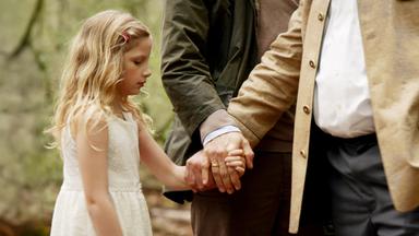 Die kleine Lucy (Carlotta von Falkenhayn) steht mit abgesenktem Blick in einem weißen Kleid im Wald und hält die Hand eines korpulenten Mannes.