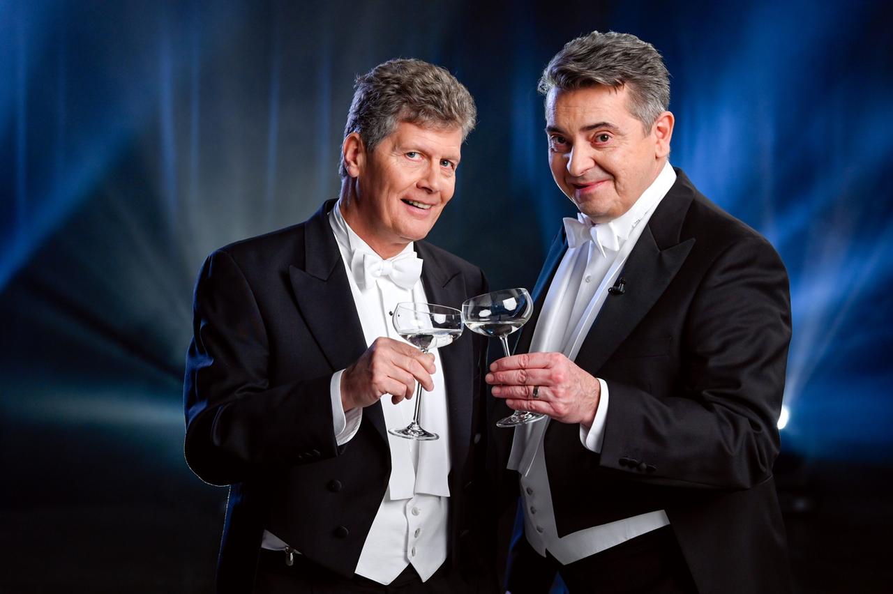 Das Bild zeigt zwei Männer in formeller Abendgarderobe, die in einem festlichen Setting stehen. Beide tragen schwarze Anzüge mit weißen Hemden und Fliegen. Die Männer halten in ihren Händen jeweils ein Glas, das mit einem klaren Getränk gefüllt ist.   Der Hintergrund ist sanft beleuchtet, mit blauen und weißen Lichtern, die eine festliche Atmosphäre schaffen. Die Männer stehen sich leicht zugewandt und scheinen freundlich zu lächeln, was eine einladende Stimmung vermittelt.   Der gesamte Rahmen des Bildes vermittelt eine elegante und feierliche Stimmung, passend zu einem Opernball-Ereignis.