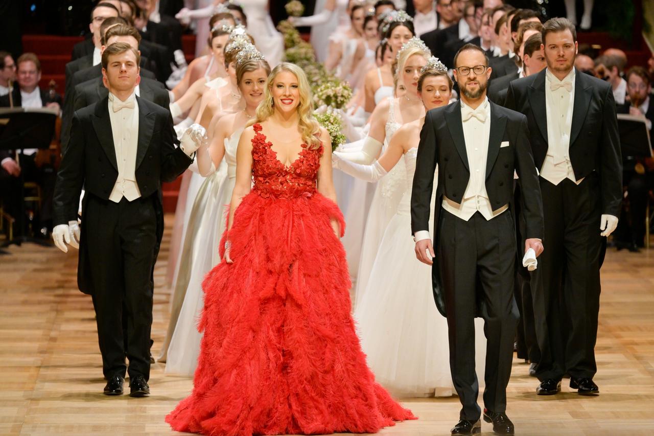 Das Bild zeigt eine festliche Szene im Ballsaal der Wiener Staatsoper, vermutlich während des Wiener Opernballs 2023. Im Vordergrund befinden sich mehrere Personen, die in einer Reihe stehen. Eine Frau in einem auffälligen, langen, roten Kleid mit Federdetails lächelt nach vorne. Neben ihr steht ein Mann in einem eleganten weißen Frack. Hinter ihnen sind zahlreiche weitere Personen zu sehen, die in weißen Abendkleidern und mit Handschuhen gekleidet sind. Einige tragen Kronen oder andere festliche Accessoires.  Der Boden des Ballsaals ist aus hellem Holz und verleiht der Szene eine feierliche Atmosphäre. Im Hintergrund ist eine Gruppe von Musikern zu sehen, die anscheinend ein Orchester bilden und für die musikalische Begleitung sorgen. Die gesamte Szene vermittelt Eindruck von Eleganz und Feierlichkeit.