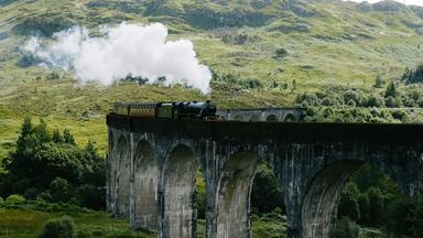 Historischer Dampfzug fährt über ein Steinviadukt in einer grünen Hügellandschaft der schottischen Highlands.