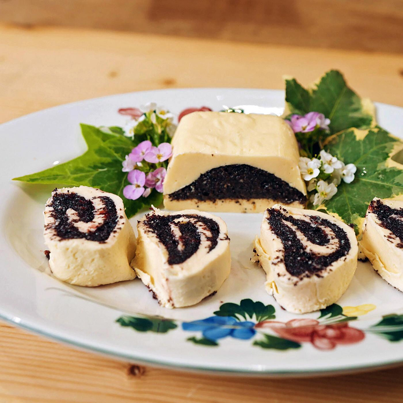 Das Bild zeigt eine auf einem weißen Teller angerichtete Speise. Der Teller hat eine dekorative Blütenmusterung am Rand. Auf dem Teller sind mehrere Stücke einer begefüllten Mohnbutter sichtbar. Diese sind in Form von kleinen, spiralförmigen Rollen geschnitten, die gelbe Füllung und eine dunkle Mohnfüllung aufweisen. In der Mitte des Tellers befindet sich ein größeres Stück der Mohnbutter, das ebenfalls in der Form eines Rechtecks präsentiert wird.   Um die Mohnbutter sind vereinzelte grüne Blätter und kleine blühende Pflanzen angeordnet, was dem Arrangement eine frische Note verleiht. Der Untergrund ist eine helle Holzfläche, auf der der Teller platziert ist.