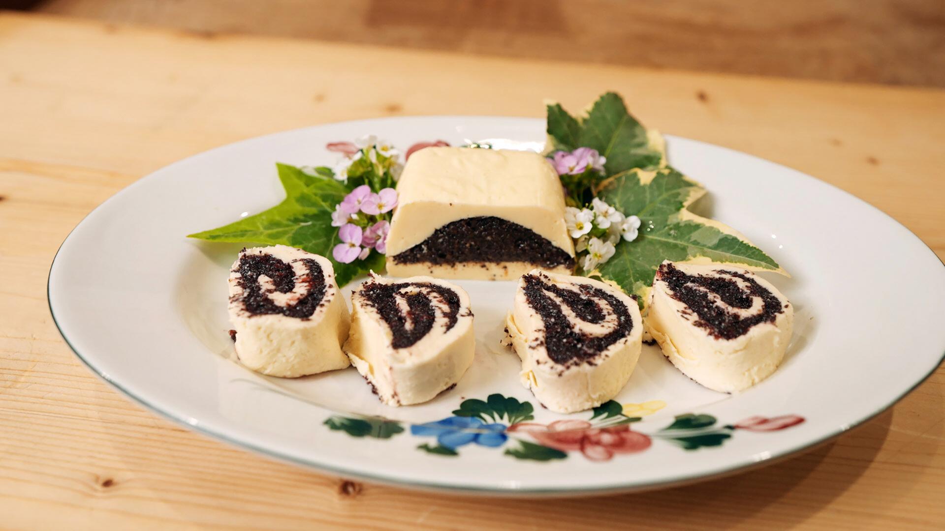 Das Bild zeigt eine auf einem weißen Teller angerichtete Speise. Der Teller hat eine dekorative Blütenmusterung am Rand. Auf dem Teller sind mehrere Stücke einer begefüllten Mohnbutter sichtbar. Diese sind in Form von kleinen, spiralförmigen Rollen geschnitten, die gelbe Füllung und eine dunkle Mohnfüllung aufweisen. In der Mitte des Tellers befindet sich ein größeres Stück der Mohnbutter, das ebenfalls in der Form eines Rechtecks präsentiert wird.   Um die Mohnbutter sind vereinzelte grüne Blätter und kleine blühende Pflanzen angeordnet, was dem Arrangement eine frische Note verleiht. Der Untergrund ist eine helle Holzfläche, auf der der Teller platziert ist.