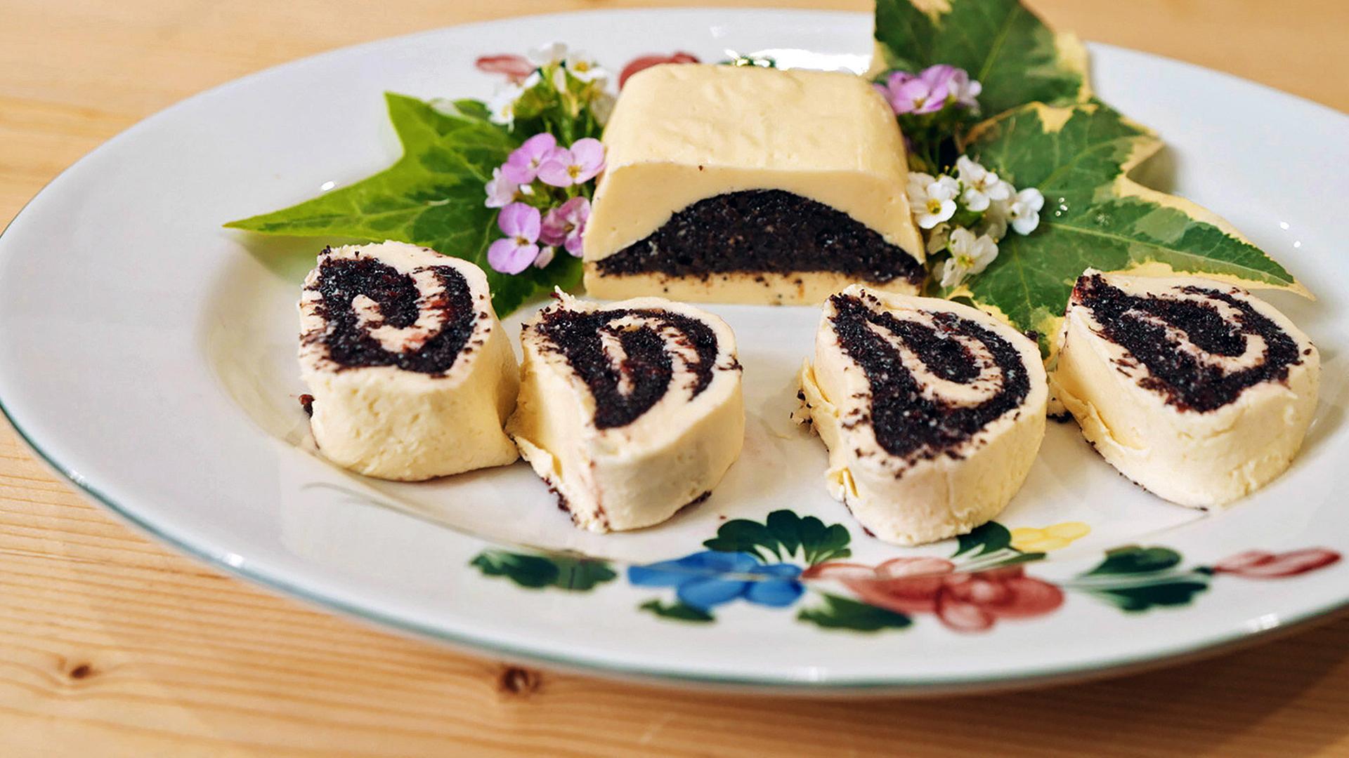 Das Bild zeigt eine auf einem weißen Teller angerichtete Speise. Der Teller hat eine dekorative Blütenmusterung am Rand. Auf dem Teller sind mehrere Stücke einer begefüllten Mohnbutter sichtbar. Diese sind in Form von kleinen, spiralförmigen Rollen geschnitten, die gelbe Füllung und eine dunkle Mohnfüllung aufweisen. In der Mitte des Tellers befindet sich ein größeres Stück der Mohnbutter, das ebenfalls in der Form eines Rechtecks präsentiert wird.   Um die Mohnbutter sind vereinzelte grüne Blätter und kleine blühende Pflanzen angeordnet, was dem Arrangement eine frische Note verleiht. Der Untergrund ist eine helle Holzfläche, auf der der Teller platziert ist.