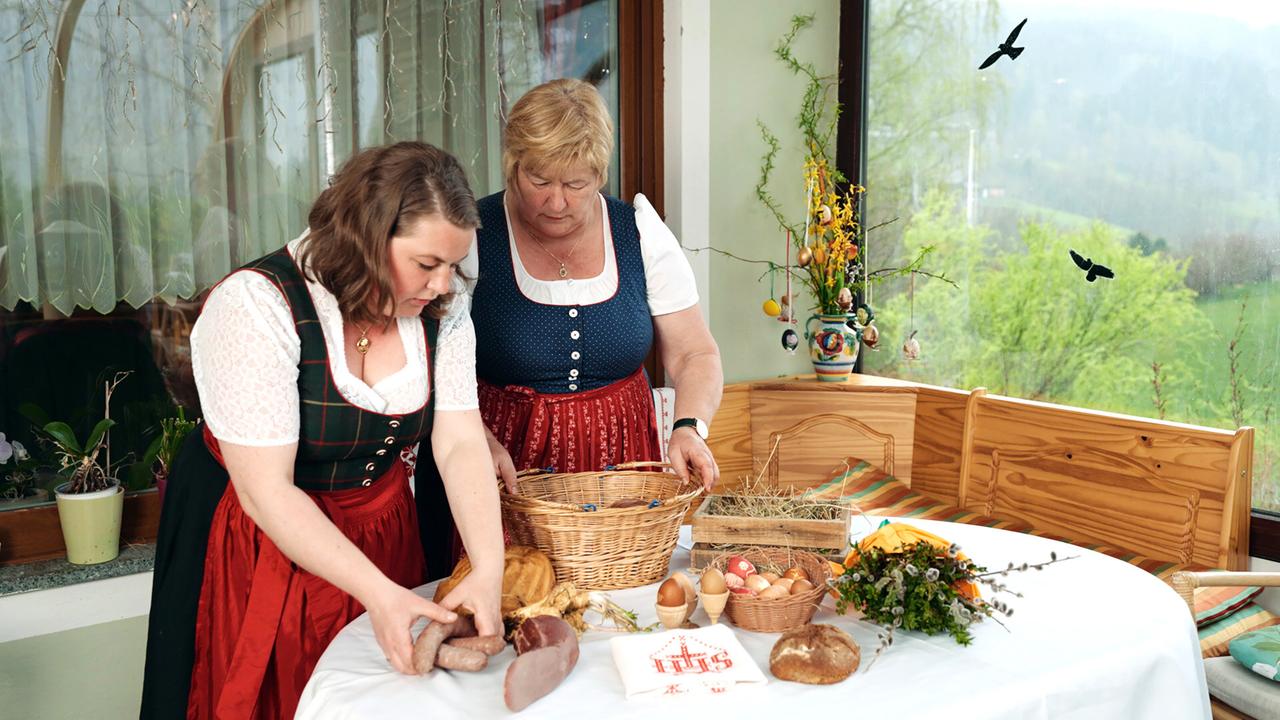 Das Bild zeigt zwei Frauen, die in einer ländlichen Umgebung aktiv sind. Sie stehen an einem Tisch, der mit einer weißen Tischdecke bedeckt ist. Auf dem Tisch sind verschiedene Nahrungsmittel und Gegenstände für die Osterjause angeordnet, darunter:  - Ein Korb aus Weidenruten, der mit blauen Eiern gefüllt ist. - Eine Holzkiste mit Heu. - Mehrere Oster- und Hühnereier, einige mit Muster. - Eine Auswahl an Lebensmitteln, darunter Wurstwaren, die auf dem Tisch liegen. - Ein Brot und ein weiterer, schmackhaft aussehender Laib, das auf der Tischdecke platziert ist. - Eine kleine, florale Dekoration aus frischen Zweigen.  Die beiden Frauen tragen traditionelle österreichische Trachten. Eine von ihnen hat lange, lockige Haare, die andere ist blond und trägt die Tracht über einem weißen Blusenausschnitt. Die Umgebung ist hell erleuchtet, und durch das Fenster ist eine grüne, hügelige Landschaft sichtbar. Auf der Fensterbank stehen Pflanzen.  Im Hintergrund sind Fenster mit Vorhängen zu sehen, die aus hellem Stoff gefertigt sind. Über dem Tisch hängt eine dezente Osterdekoration, und die Atmosphäre wirkt festlich und gemeinschaftlich.