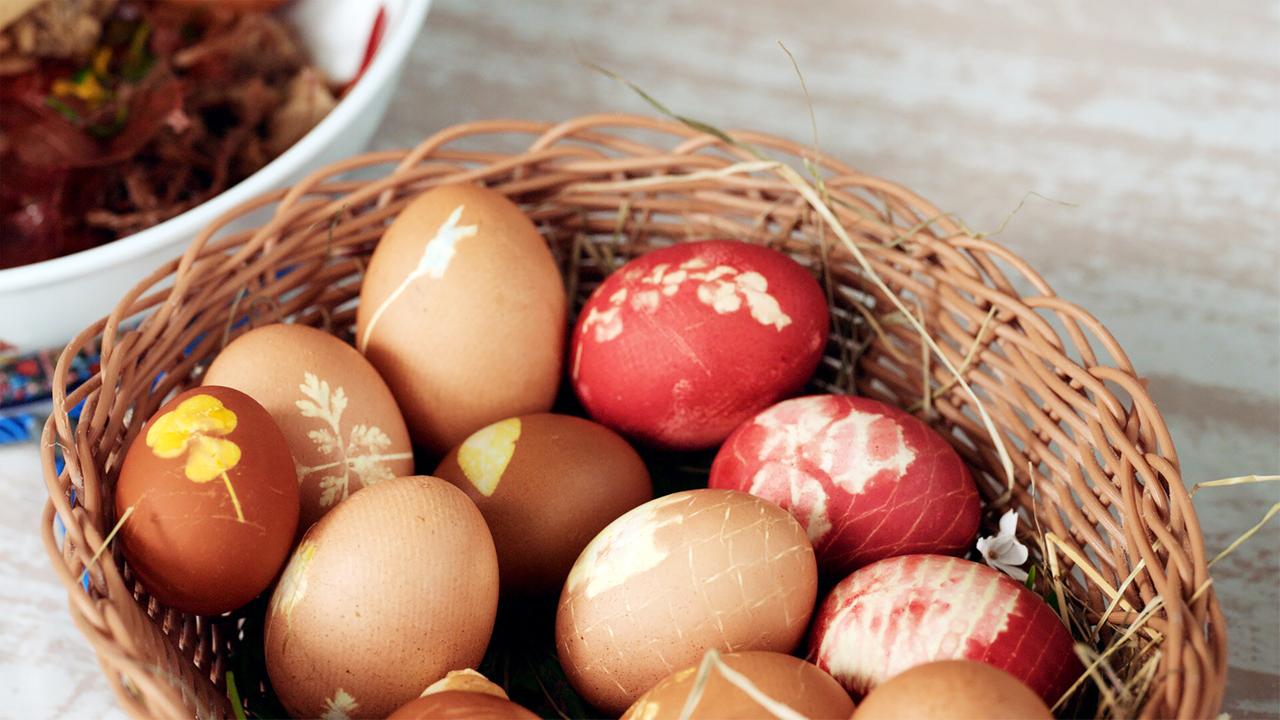 Das Bild zeigt einen Korb mit natürlich gefärbten Ostereiern. Der Korb ist geflochten und aus braunem Weidengeflecht. Die Eier sind unterschiedlich gefärbt: Einige sind in sanften Erdtönen gehalten, während andere ein kräftiges Rot aufweisen. Die Eier sind teilweise mit Pflanzenmustern verziert, die durch die Färbetechnik sichtbar gemacht wurden.   Im Hintergrund ist eine weiße Schüssel zu sehen, die auf einer Holzoberfläche steht, die eine helle, leicht strukturiert wirkende Farbgebung hat. Auf dem Tisch liegen einige grüne Halme und kleine Blüten, die einen Bezug zu Ostern und der Natur herstellen. Die gesamte Komposition vermittelt eine festliche und gemütliche Atmosphäre, die mit dem Brauch der Ostereierfärbung verbunden ist.