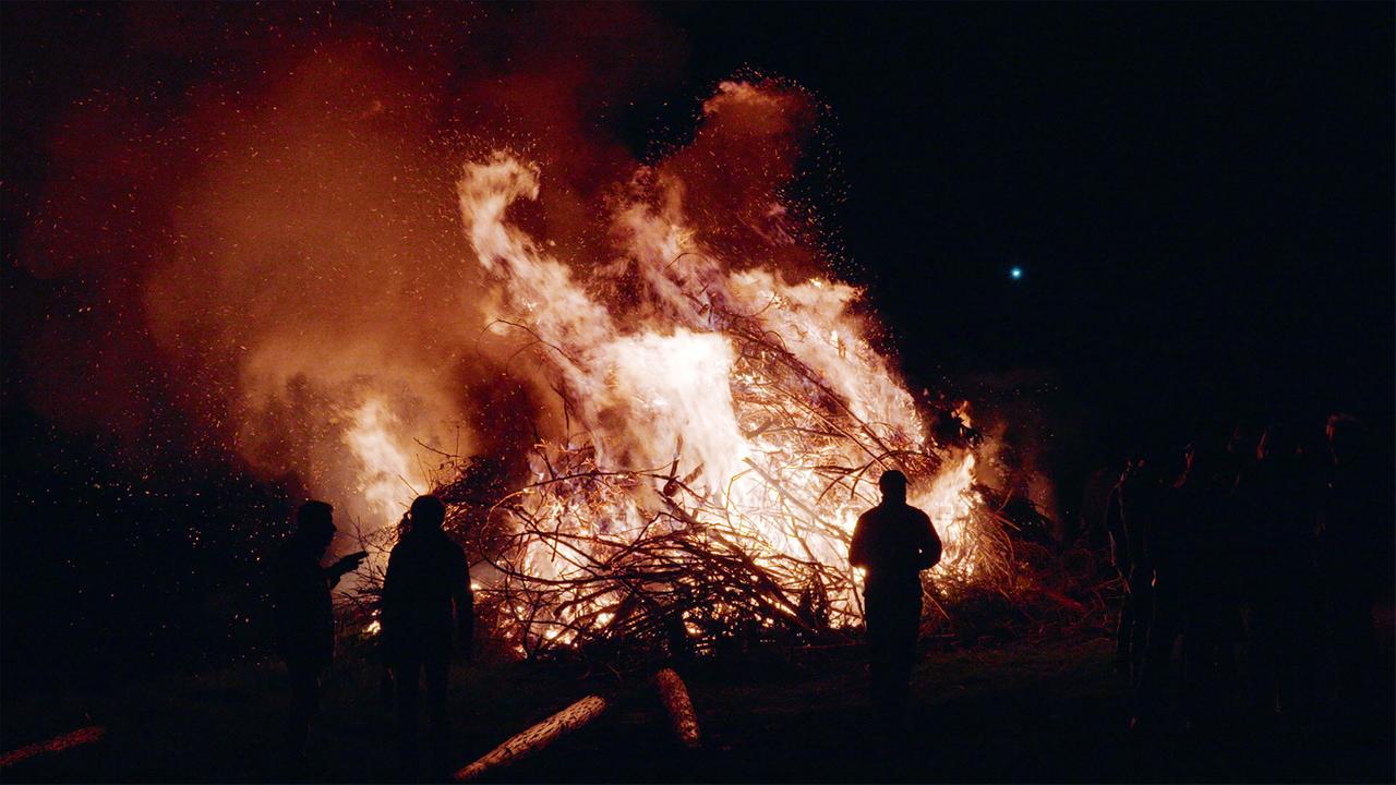 Die Darstellung zeigt ein großes Osterfeuer, das in der Dunkelheit brennt. Flammen leuchten hell und erzeugen eine orangefarbene und goldene Farbenpracht, während Rauch und Funken in die Luft aufsteigen. Um das Feuer versammeln sich mehrere Menschen, die als dunkle Silhouetten zu erkennen sind. Die Konturen der Personen sind unscharf, und sie scheinen zu beobachten oder miteinander zu sprechen. Der Hintergrund ist dunkel, was den Fokus auf das Feuer und die umstehenden Menschen lenkt. Es herrscht eine festliche Atmosphäre, die durch das brennende Osterfeuer und die Versammlung von Gemeinschaftsmitgliedern symbolisiert wird.