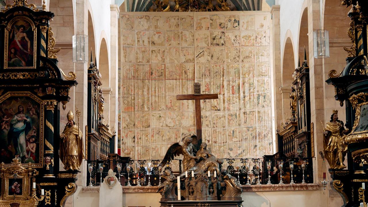 Das Bild zeigt das innere einer Kirche, wahrscheinlich in Gurk, Österreich. Im Hintergrund ist ein großes Fastentuch zu sehen, das mit vielen farbigen Bildern bedruckt ist und eine religiöse Erzählung darstellt. Darüber befindet sich ein hölzernes Kreuz, das im Vordergrund sichtbar ist.   Auf beiden Seiten des Kreuzes sind kunstvoll gestaltete, goldene Statuen dargestellt, die Heilige oder Engel darstellen könnten. Im unteren Bereich des Altares sind Kerzenständer und weitere detailreiche Elemente aus Stein und Holz zu erkennen. Die Wände der Kirche sind aus hellem Stein, und die Decke zeigt farbige Balken, die der Szene eine festliche Atmosphäre verleihen.   Das Bild vermittelt einen Eindruck von religiösem Brauchtum und Gemeinschaft, insbesondere in der Osterzeit.