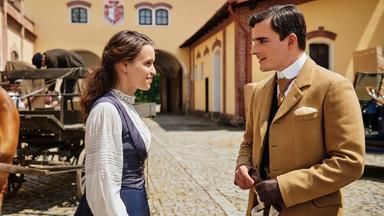 Eine junge Frau und ein junger Mann in historischer Kleidung stehen neben einer Pferdekutsche und lächeln sich freundlich an.