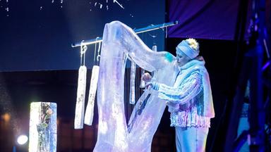 Eine Frau in silbern glitzernder Kleidung spielt eine Harfe aus Eis, im Hintergrund hängt ein Glockenspiel aus Eis.