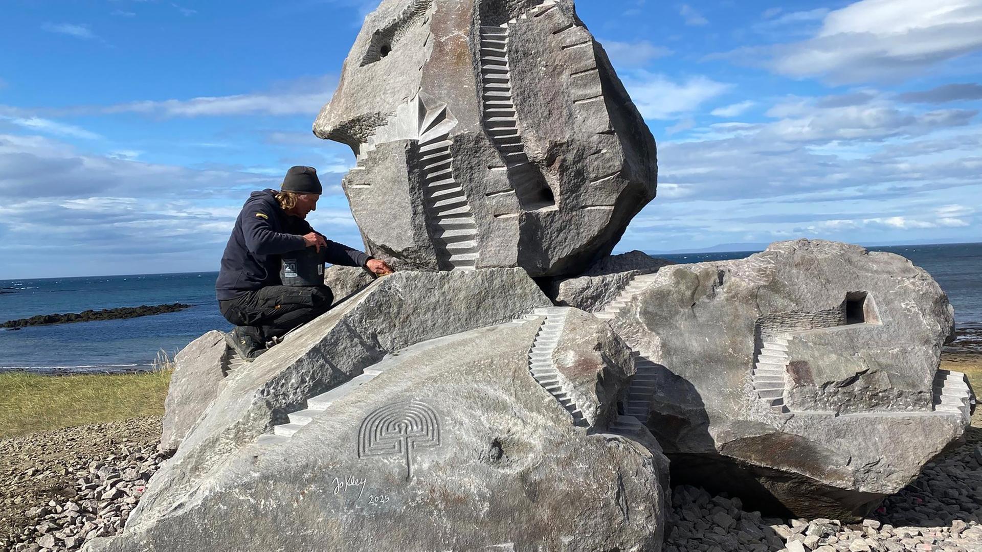 Jo Kley arbeitet an einem Felsen am Meer, der mit eingemeißelten Stufen, Petroglyphen und einem Gesicht zur Skulptur gestaltet ist.