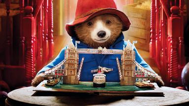 Paddington steht vor dem aufgeschlagenen Klapp-Bilderbuch und schaut sich voller Bewunderung ein Modell der "Tower Bridge" an.
