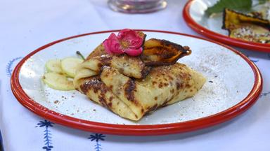 Auf dem Bild ist ein Teller mit Palatschinken zu sehen, die auf einer weißen Unterlage mit blauen Mustern platziert sind. Die Palatschinken sind gefüllt und leicht angeröstet, mit einer goldbraunen und teilweise knusprigen Oberfläche. Auf den Palatschinken ruht eine dekorative pinke Blüte. Neben den Palatschinken liegen schmale Scheiben von äpfeln, und der Teller ist von einem roten Rand umgeben. Im Hintergrund sind weitere Teller zu erkennen, die ebenfalls Palatschinken oder ähnliches enthalten. Es wird eine rustikale und ansprechende Atmosphäre vermittelt, die an eine traditionelle österreichische Gastlichkeit erinnert.