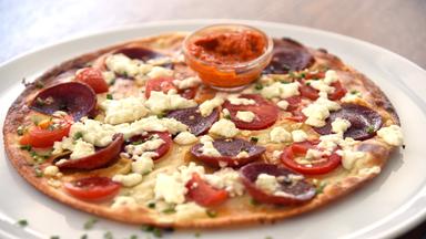 Das Bild zeigt einen flachen, runden Pfannkuchen, der auf einem weißen, runden Teller angerichtet ist. Der Pfannkuchen ist leicht gebräunt und hat eine goldene Farbe. Er ist mit verschiedenen Zutaten belegt: dünne Scheiben von Tomaten, leicht geräucherter Wurst, die laut Beschreibung eine türkische Knoblauchwurst sein soll, sowie Stücke von Schafskäse, die unregelmäßig auf der Oberfläche verteilt sind.   In der Mitte des Pfannkuchens befindet sich eine kleine, durchsichtige Schale mit einer roten Sauce, die möglicherweise eine Paprika- oder Chilisauce ist. Darüber wurden frische, gehackte Schnittlauchstücke gestreut, die dem Gericht Farbe und Aroma verleihen. Der Hintergrund ist unscharf, was den Fokus auf den Pfannkuchen lenkt.