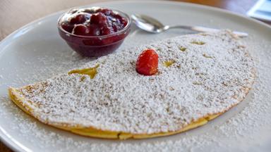 Das Bild zeigt einen Teller mit einem großen, gefalteten Pfannkuchen, der mit Puderzucker bestäubt ist. In der Mitte des Pfannkuchens liegt eine kleine Erdbeere als Dekoration. Neben dem Pfannkuchen befindet sich eine kleine Schüssel mit einer dunklen, glatten Fruchtsoße, die auf Kirschen oder eine ähnliche Frucht hindeutet. Der Teller ist schlicht und hat einen weißen Rand, der eine neutrale, helle Tischoberfläche kontrastiert. Die gesamte Präsentation wirkt appetitlich und einladend.