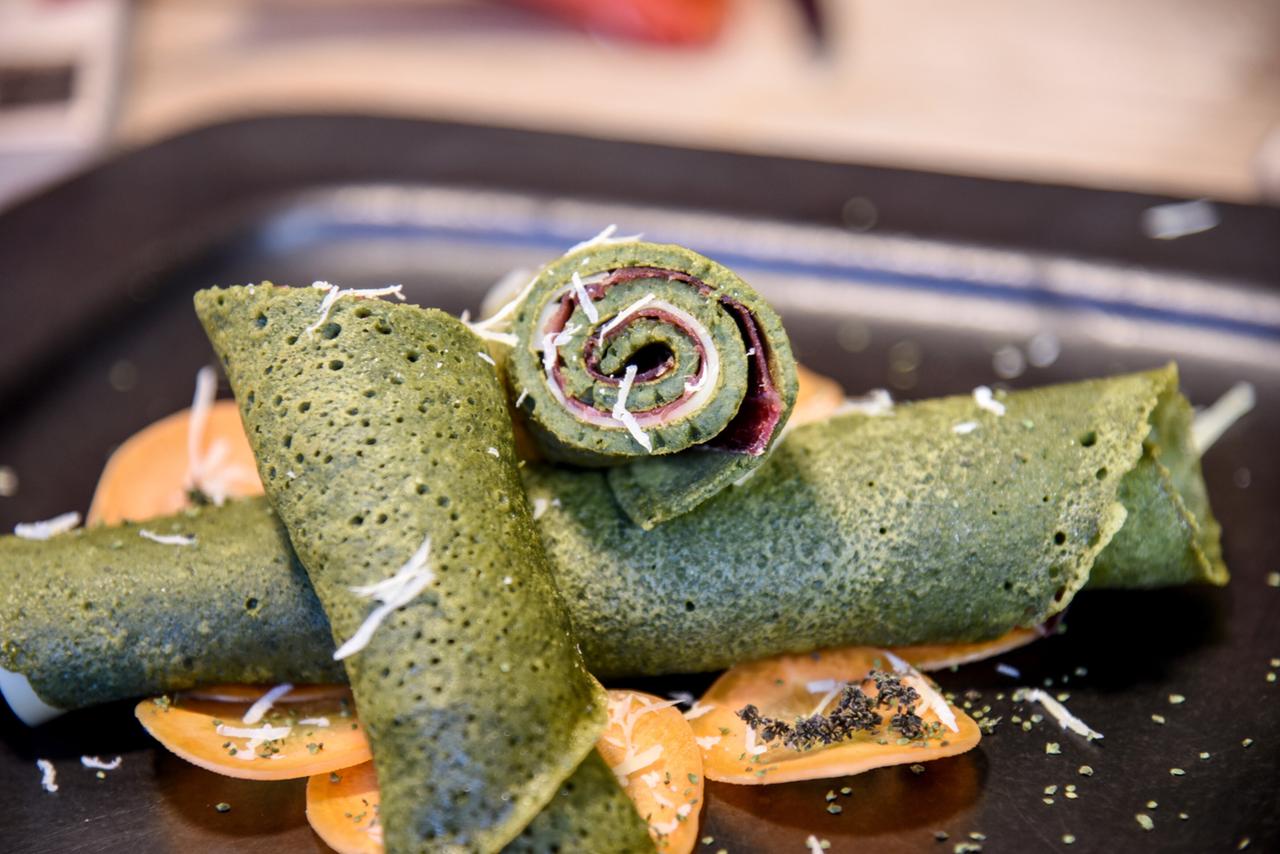 Das Bild zeigt eine Auswahl von grünen Palatschinken, die auf einem schwarzen Teller angerichtet sind. Zwei flache, grüne Teigfladen liegen horizontal auf dem Teller, während ein dritter Teigfladen aufgerollt im Vordergrund platziert ist. Die Palatschinken scheinen mit einer Füllung versehen zu sein, möglicherweise mit einer Art Wurst oder Käse, basierend auf der Sichtbarkeit von rosanen und weißen Schichten im aufgerollten Stück. 

Unter den Palatschinken sind orangefarbene Scheiben, möglicherweise von einer Frucht oder einer Gemüsesorte, die als Dekoration dienen. Auf den grünen Teigfladen liegt etwas geriebener Käse oder ein ähnliches heller, schuppiges Topping. Die Präsentation ist ordentlich, und das Gesamtbild vermittelt einen ansprechenden Eindruck von einem kreativen Gericht.