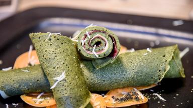 Das Bild zeigt eine Auswahl von grünen Palatschinken, die auf einem schwarzen Teller angerichtet sind. Zwei flache, grüne Teigfladen liegen horizontal auf dem Teller, während ein dritter Teigfladen aufgerollt im Vordergrund platziert ist. Die Palatschinken scheinen mit einer Füllung versehen zu sein, möglicherweise mit einer Art Wurst oder Käse, basierend auf der Sichtbarkeit von rosanen und weißen Schichten im aufgerollten Stück. 

Unter den Palatschinken sind orangefarbene Scheiben, möglicherweise von einer Frucht oder einer Gemüsesorte, die als Dekoration dienen. Auf den grünen Teigfladen liegt etwas geriebener Käse oder ein ähnliches heller, schuppiges Topping. Die Präsentation ist ordentlich, und das Gesamtbild vermittelt einen ansprechenden Eindruck von einem kreativen Gericht.