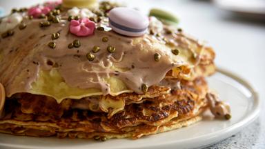 Das Bild zeigt eine große, mehrlagige Palatschinkentorte, die auf einem runden weißen Teller angerichtet ist. Die Torte besteht aus mehreren dünnen, goldbraun gebratenen Teigschichten, die übereinander gestapelt sind. Die oberste Schicht ist mit einer cremigen, hellbraunen Füllung bedeckt, die einen glatten, glänzenden Überzug hat.  Die Torte ist mit bunten Dekorationen verziert. Darunter befinden sich kleine, runde, pastellfarbene Makronen und bunte Zuckerstreusel in verschiedenen Größen. Es sind auch essbare Blüten und kleine, glänzende, goldene Perlen zu sehen, die für zusätzliches Farbspiel und Textur sorgen.  Der Hintergrund ist unscharf, was die Aufmerksamkeit auf die Torte lenkt. Die gesamte Präsentation vermittelt einen ansprechenden, einladenden Eindruck und wirkt sehr appetitlich.