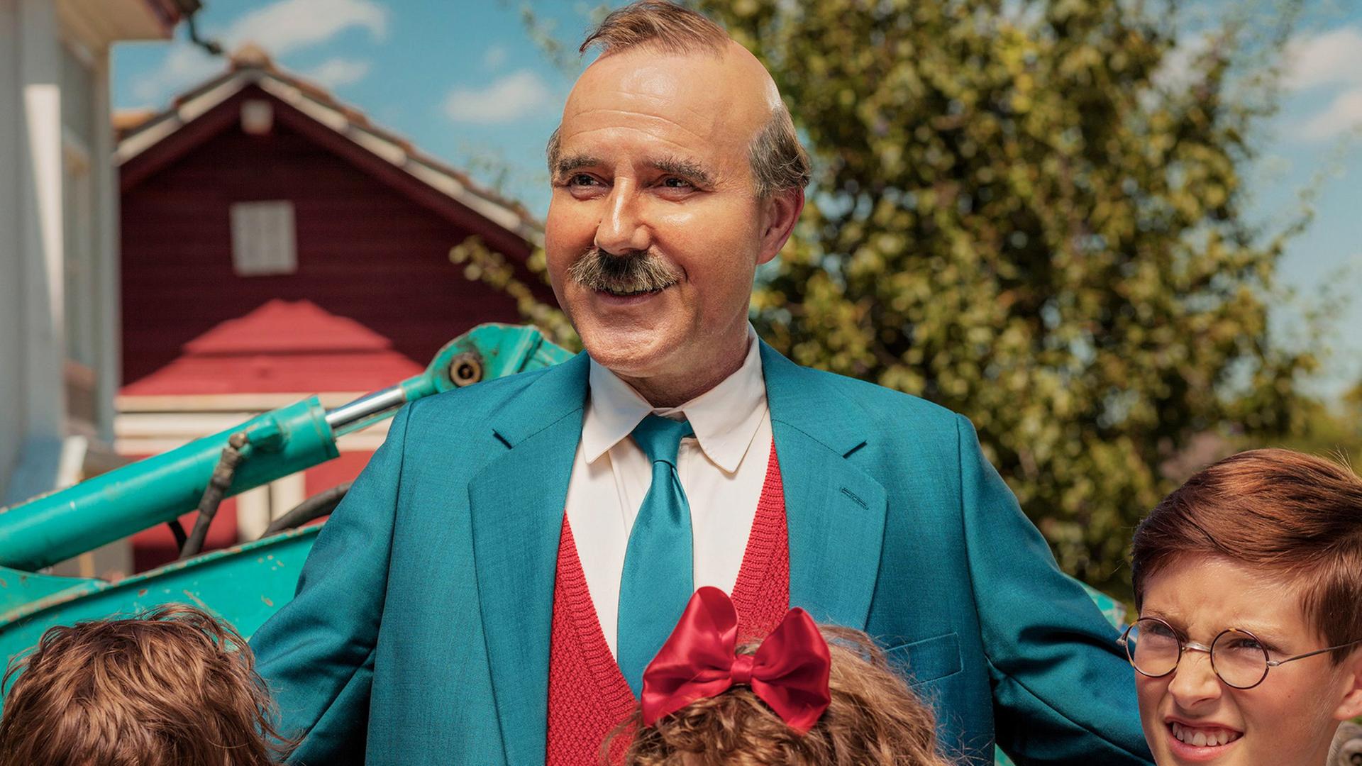 Stefan Kurt als Papa Moll steht lächelnd mit seinen Kindern. Links ist ein Junge mit braunen Haaren und ernstem Gesichtsausdruck, in der Mitte ein Mädchen mit lockigen Haaren und einer großen roten Schleife, rechts ein Junge mit Brille und braunen Haaren. Im Hintergrund sind ein grüner Bagger, ein rotes Gebäude und Bäume zu sehen.