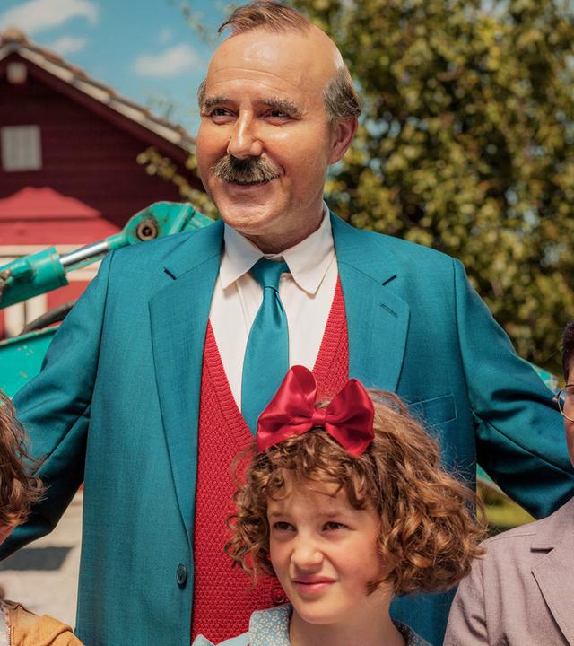 Stefan Kurt als Papa Moll steht lächelnd mit seinen Kindern. Links ist ein Junge mit braunen Haaren und ernstem Gesichtsausdruck, in der Mitte ein Mädchen mit lockigen Haaren und einer großen roten Schleife, rechts ein Junge mit Brille und braunen Haaren. Im Hintergrund sind ein grüner Bagger, ein rotes Gebäude und Bäume zu sehen.