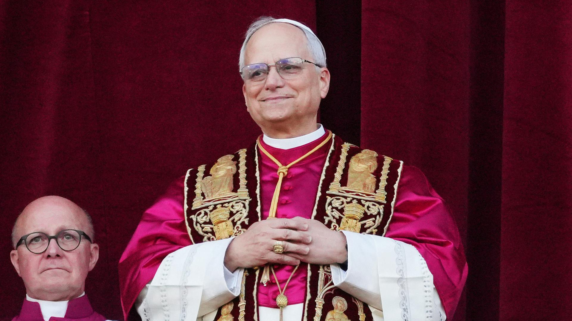 Mann im Papst-Ornat steht lächelnd vor einem purpurnen Vorhang.