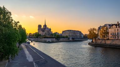 Blick über die Seine auf die Kathedrale Notre-Dame in Paris. Die untergehende Sonne taucht den Himmel hinter der Île de la Cité in warmes Licht.