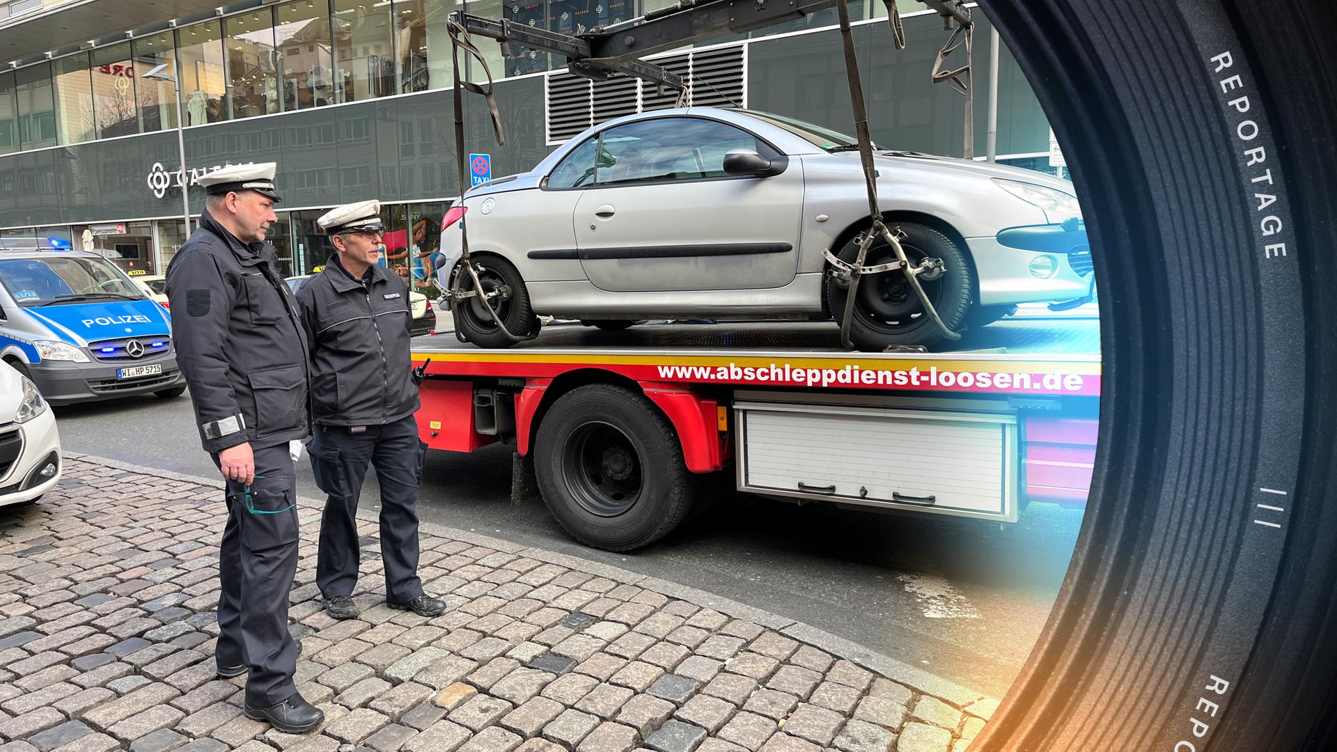 Ein silbernes Auto wurde gerade auf einen Abschleppwagen geladen, es ist noch am Kran befestigt. Links daneben stehen Stephan Janisch und Rainer Deckert von der städtischen Verkehrspolizei Frankfurt und überwachen den Vorgang.