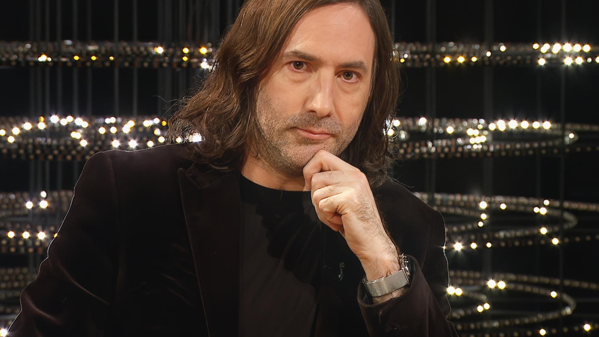 Ein Mann mit mittellangen braunen Haaren und dunklem Jackett sitzt nachdenklich mit der Hand am Kinn vor einer Kulisse mit vielen kleinen Lichtern.