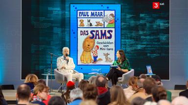 Ein älterer Mann und eine Frau sitzen auf einer Bühne in Sesseln und unterhalten sich vor Publikum. Im Hintergrund ist ein Kinderbuch eingeblendet.