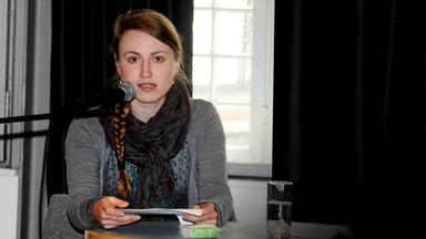 Eine junge Frau sitzt an einem Tisch und liest von einem Blatt laut in ein Mikrofon.