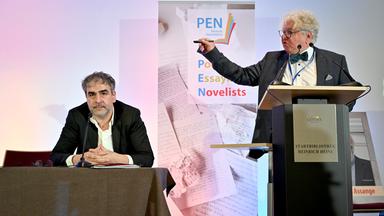 Der PEN--Vorsitzende Deniz Yücel sitzt bei der Pen-Mitgliederversammlung an einem Podest, während PEN-Mitglied Christoph Nix neben ihm an einem Podest wild gestikulierend redet.