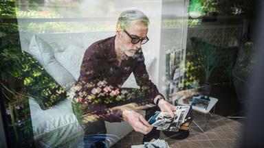 Blick durch eine spiegelnde Scheibe: Ein Mann mit geraumeliertem Haar, Bart und dunkler Brille sitzt auf einem Sofa und betrachtet Fotos.