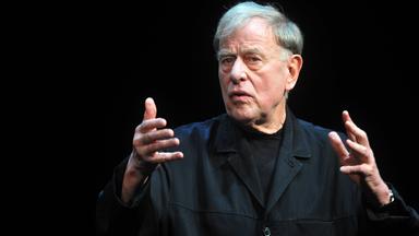 Claus Peymann steht vor schwarzem Hintergrund auf einer Bühne, trägt einen schwarzen Mantel und gestikuliert mit ausgebreiteten Händen.