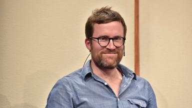 Ein Mann mit kurzen Haaren, Bart und Brille, hat ein kleines Mikro an die Wange geklemmt und lächelt in die Kamera.