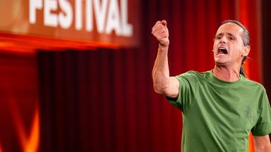 Philipp Weber steht auf der rechten Seite des Bildes. Mit seiner rechten Hand deutet er in Richtung des linken Bildrandes. Er trägt ein grünes T-Shirt. Der Hintergrund ist rot