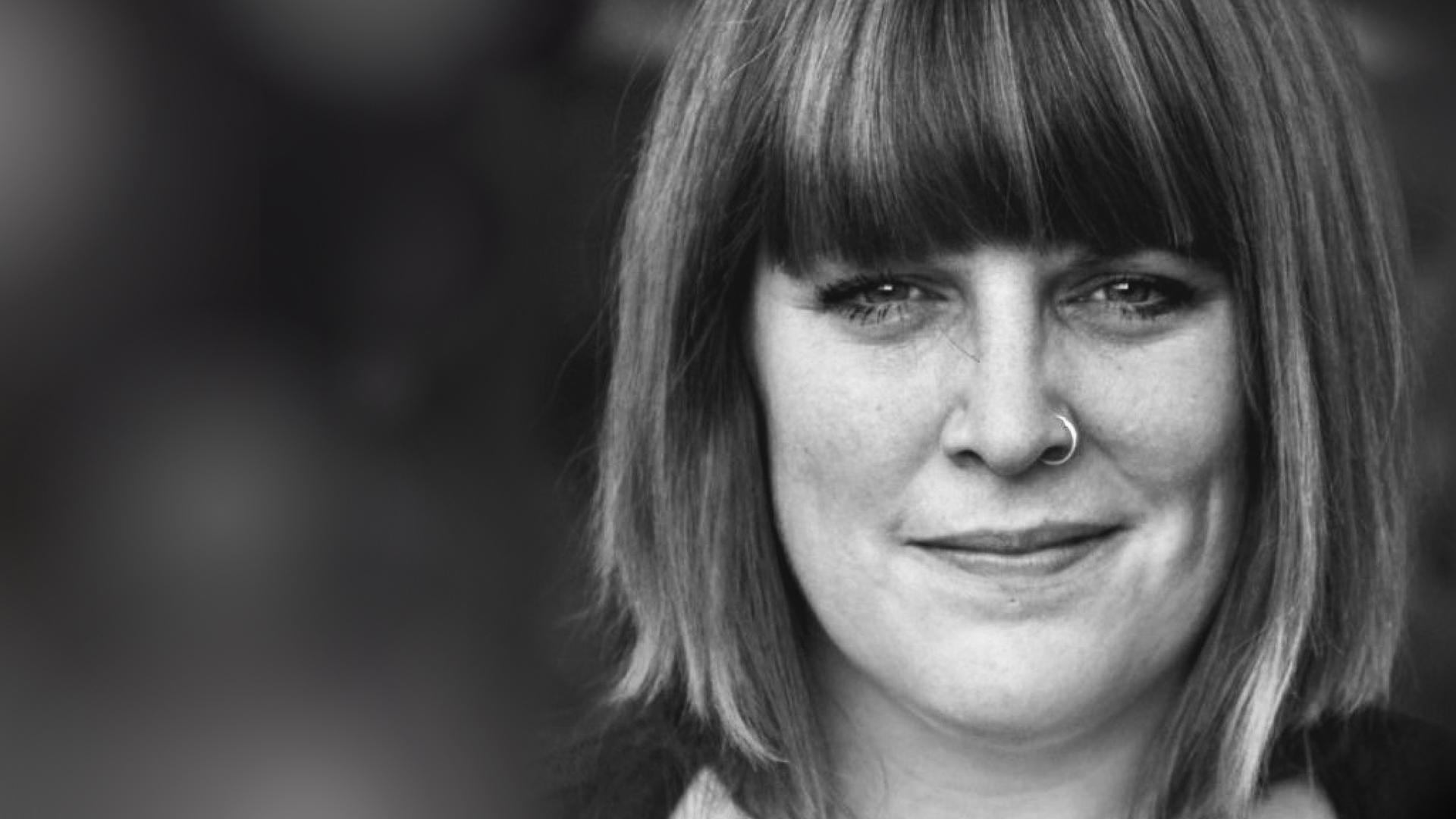 Das Gesicht einer Frau mit Pony, schulterlangem Haar und Nasenpiercing. 